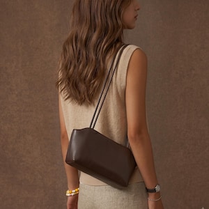 May include: A dark brown rectangular shoulder bag with a smooth finish and two thin, long straps. The bag features a structured, elongated horizontal shape and is worn over the shoulder, resting against the side of the torso.