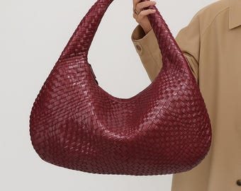 May include: A burgundy woven leather shoulder bag with a large, curved handle. The bag has a rounded shape and a textured surface, showcasing a detailed weave pattern. The bag is held by a person wearing a tan jacket.
