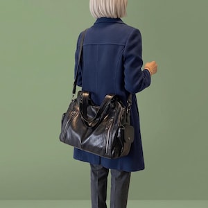 May include: A black leather-look shoulder bag featuring multiple exterior zippered pockets, dual top handles, and an adjustable crossbody strap. The bag has a structured rectangular shape with silver-toned hardware accents and side compartments.