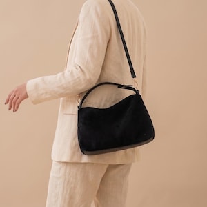 May include: A black suede handbag with a curved top handle and a long, thin, adjustable black shoulder strap. The bag features a dark brown base and gold-toned metal hardware attachments for the straps, providing a structured yet soft appearance.