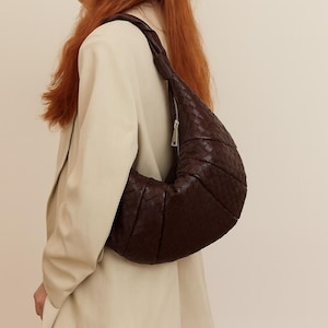 May include: A dark brown, woven leather shoulder bag with a crescent shape. The bag features a zipper closure and a braided strap. The bag is worn over the shoulder.