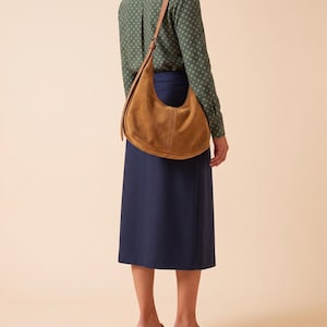 May include: A tan suede hobo bag with a long shoulder strap, worn over a green long-sleeve shirt with a small white geometric pattern, a solid navy blue midi skirt, and tan woven heeled mules. The bag features a curved shape and visible stitching.