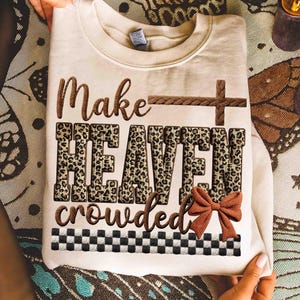 May include: Cream-colored sweatshirt with the text "Make HEAVEN crowded" in brown and leopard print. A brown cross and bow accent the design. A black and white checkered pattern is at the bottom.
