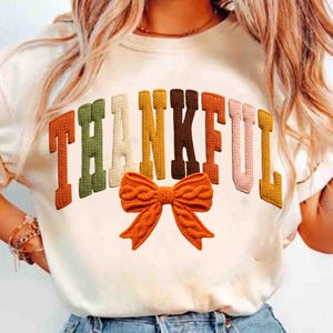 May include: A cream-coloured t-shirt featuring the word "THANKFUL" in textured, multi-coloured letters. An orange knitted bow is positioned below the word. The letters are in shades of orange, green, yellow, brown, pink, and cream.