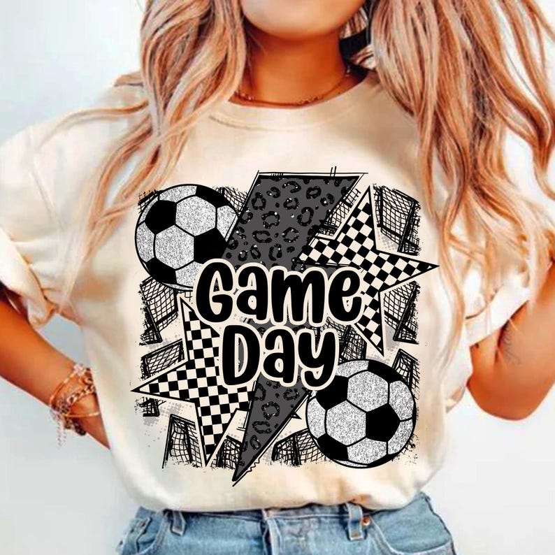 May include: White t-shirt with a black and white graphic design featuring two soccer balls, a checkered star, a leopard print lightning bolt, and the text "Game Day".