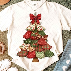 Peut inclure: T-shirt blanc avec un motif de sapin de Noël. L'arbre est composé de formes de tissu colorées superposées en rouge, vert et imprimés à motifs, ornées de nœuds rouges. Le t-shirt a un col rond et des manches courtes.