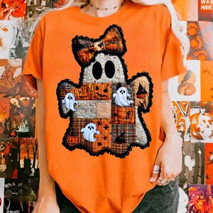 May include: Orange t-shirt with a patchwork ghost design, a plaid bow, pumpkins, and ghost appliqués. The ghost has black eyes and is trimmed with black lace. A Halloween-themed graphic tee.