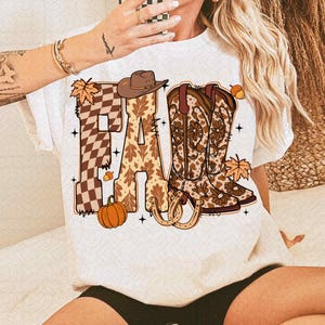 May include: White t-shirt with the word "FALL" in a fall-themed design. The "F" is a brown and tan checkered pattern, the "A" has a leaf pattern, and the "LL" features cowboy boots. A pumpkin, horseshoe, and leaves complete the design.
