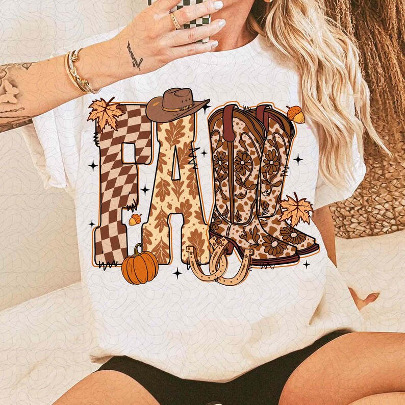 May include: White t-shirt with the word "FALL" in a fall-themed design. The "F" is a brown and tan checkered pattern, the "A" has a leaf pattern, and the "LL" features cowboy boots. A pumpkin, horseshoe, and leaves complete the design.