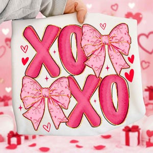May include: White pillow with a pink "XOXO" design. The letters are large and bubble-style, with gold outlines. Two pink bows with bow patterns are featured, along with red hearts and stars. A Valentine's Day theme.