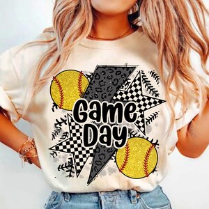 Op de afbeelding: Cr&egrave;mekleurig T-shirt met een "Game Day"-afbeelding. Het ontwerp toont twee gele softballs met rode stiksels, een zwart-wit geblokte ster, een grijze bliksemschicht met luipaardprint en honkbalstiksels.