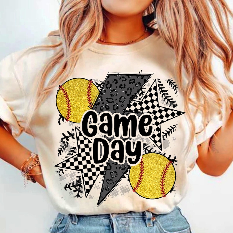 Op de afbeelding: Cr&egrave;mekleurig T-shirt met een "Game Day"-afbeelding. Het ontwerp toont twee gele softballs met rode stiksels, een zwart-wit geblokte ster, een grijze bliksemschicht met luipaardprint en honkbalstiksels.