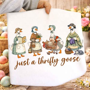 Könnte beinhalten: Weißer Stoff mit einer Illustration von vier Gänsen in Vintage-Kleidung. Jede Gans wird mit verschiedenen Accessoires und Aktivitäten dargestellt. Der Text "just a thrifty goose" ist in brauner Schrift unten geschrieben.