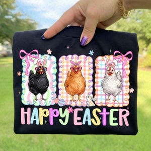 May include: Black t-shirt with a colorful "Happy Easter" graphic. The graphic features three chickens wearing bunny ears and sunglasses, set against a checkered background with pink bows and flowers. A cute Easter design.