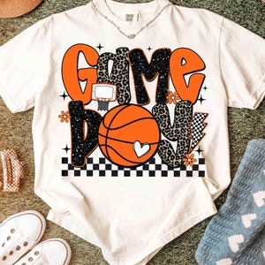 Peut inclure: T-shirt cr&egrave;me avec un motif sur le th&egrave;me du basketball. Le motif pr&eacute;sente les mots "GAME DAY" en orange et imprim&eacute; l&eacute;opard, un ballon de basket, un c&oelig;ur et un motif &agrave; carreaux. Le t-shirt a un col rond.
