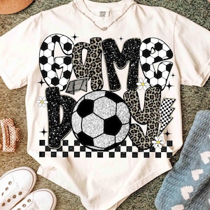 May include: Cream-colored t-shirt with a soccer-themed graphic. The design features the words "Game Day" in a mix of soccer ball patterns, leopard print, and glitter. Additional elements include a soccer net, daisies, and a checkered pattern.