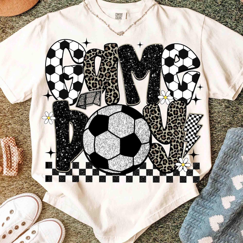 May include: Cream-colored t-shirt with a soccer-themed graphic. The design features the words "Game Day" in a mix of soccer ball patterns, leopard print, and glitter. Additional elements include a soccer net, daisies, and a checkered pattern.