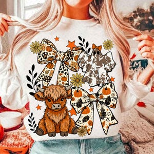 Op de afbeelding: Witte sweatshirt met een herfstthema-ontwerp. Het ontwerp bevat een bruine Schotse hooglander, een spook en twee strikken met pompoen- en bladerpatronen. Oranje sterren en groen-gele bloemen maken het herfstthema compleet.