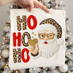 May include: White sweatshirt with a festive design featuring the text "HO HO HO" in red and leopard print. Santa Claus is winking and holding a heart. The design is embroidered with a textured, raised effect.