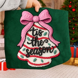 May include: Green Christmas pillow with a knitted Christmas tree design and a pink bow. The tree has red and white stripes, green leaf accents, and the phrase "tis the season" in black script.