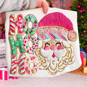Peut inclure: Coussin de Noël brodé avec un motif de Père Noël et les mots "HO HO HO". Le Père Noël porte un bonnet rose et une barbe blanche. Les lettres sont rouges, vertes et bleues avec une bordure dorée. Le coussin est tenu par une personne.