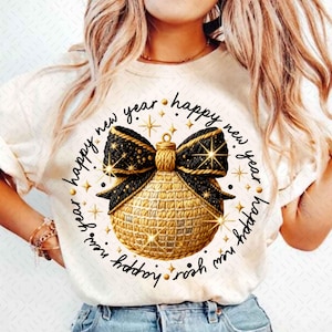 Puede incluir: Camiseta color crema con un diseño de adorno navideño dorado y negro. El adorno está rodeado por las palabras "happy new year" en un patrón circular, con acentos de estrella. Un artículo de ropa festivo.