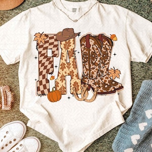 May include: A cream-coloured t-shirt with a graphic design featuring the word "FALL" in brown and tan tones. The design includes a cowboy hat, cowboy boots, a pumpkin, and autumn leaves. The shirt has a crew neck and short sleeves.