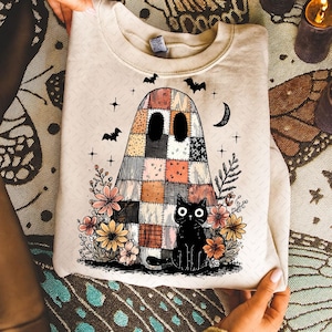 May include: Beige sweatshirt featuring a patchwork ghost design with a black cat, flowers, bats, stars, and a crescent moon. The ghost has large, dark eyes and is surrounded by colorful floral elements. A Halloween-themed graphic.