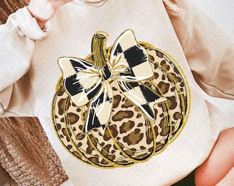 Puede incluir: Sudadera color crema con un diseño de calabaza. La calabaza tiene un estampado de leopardo con un lazo a cuadros blanco y negro. El diseño está delineado con purpurina dorada.