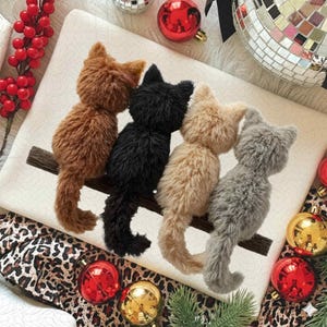 Könnte beinhalten: Vier Plüschkatzenfiguren in Braun, Schwarz, Beige und Grau, die auf einem Holzast sitzen. Die Katzen sind auf einer weißen Oberfläche angeordnet, umgeben von Weihnachtsschmuck und einer Discokugel.