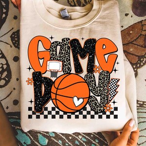 Peut inclure: Sweat-shirt cr&egrave;me avec l'inscription "Game Day Love" en orange, imprim&eacute; l&eacute;opard et noir. Le motif comprend un ballon de basket, un panier et des d&eacute;tails floraux. Un c&oelig;ur blanc est sur le ballon.