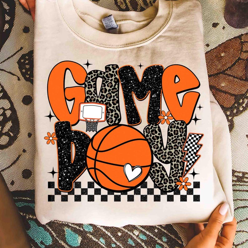 Peut inclure: Sweat-shirt cr&egrave;me avec l'inscription "Game Day Love" en orange, imprim&eacute; l&eacute;opard et noir. Le motif comprend un ballon de basket, un panier et des d&eacute;tails floraux. Un c&oelig;ur blanc est sur le ballon.