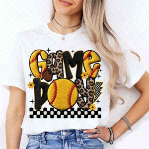 May include: White t-shirt featuring the words "GAME ON" in a playful design. The letters are a mix of yellow, leopard print, and black, with a softball graphic and lightning bolt. The design includes small flowers and a black and white checkered border.