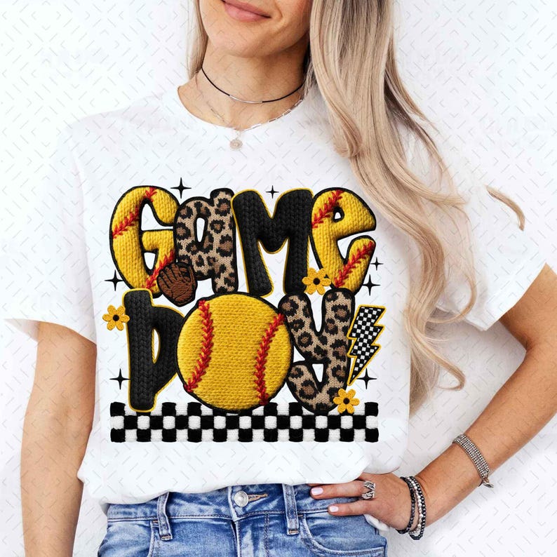 May include: White t-shirt featuring the words "GAME ON" in a playful design. The letters are a mix of yellow, leopard print, and black, with a softball graphic and lightning bolt. The design includes small flowers and a black and white checkered border.