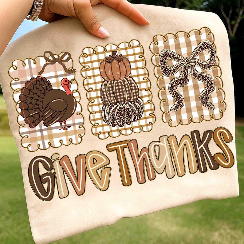 May include: A beige t-shirt featuring three decorative squares with Thanksgiving-themed designs. The squares include a turkey, stacked pumpkins, and a leopard print bow. The words "Give Thanks" are printed in a brown, tan, and white font.