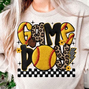 May include: A cream-colored sweatshirt featuring the words "GAME DAY" in a mix of fonts and patterns. The design includes a yellow softball, leopard print letters, and a checkered border. Additional elements include a lightning bolt and small yellow flowers.