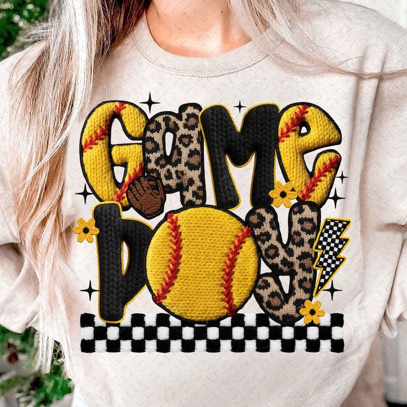 May include: A cream-colored sweatshirt featuring the words "GAME DAY" in a mix of fonts and patterns. The design includes a yellow softball, leopard print letters, and a checkered border. Additional elements include a lightning bolt and small yellow flowers.
