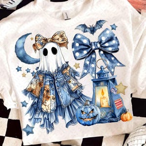 May include: White t-shirt featuring a whimsical Halloween design. The graphic includes a ghost character with a denim jacket and bow, a blue lantern, a jack-o'-lantern, and a bat. Accents include stars and a crescent moon.