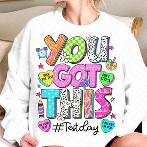 May include: White sweatshirt with a colorful graphic that says "You Got This #Testday". The graphic includes hearts, stars, butterflies, and other fun designs.