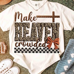 May include: White t-shirt with the text "Make HEAVEN crowded" in brown and leopard print. A brown cross and bow accent the design. The shirt has a black and white checkered pattern at the bottom.