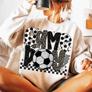 May include: Cream-colored sweatshirt with a soccer-themed graphic. The design features the words "GAME DAY" in a mix of black, leopard print, and soccer ball patterns. Includes a soccer ball, daisies, and a checkered pattern.