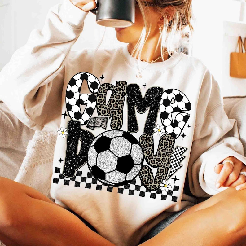 May include: Cream-colored sweatshirt with a soccer-themed graphic. The design features the words "GAME DAY" in a mix of black, leopard print, and soccer ball patterns. Includes a soccer ball, daisies, and a checkered pattern.