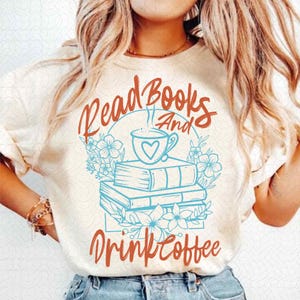 以下が含まれることがあります： クリーム色のTシャツ。テキスト「Read Books And Drink Coffee」は錆色の筆記体。デザインは本の山、ハートのコーヒーカップ、水色の花のアクセントが特徴です。