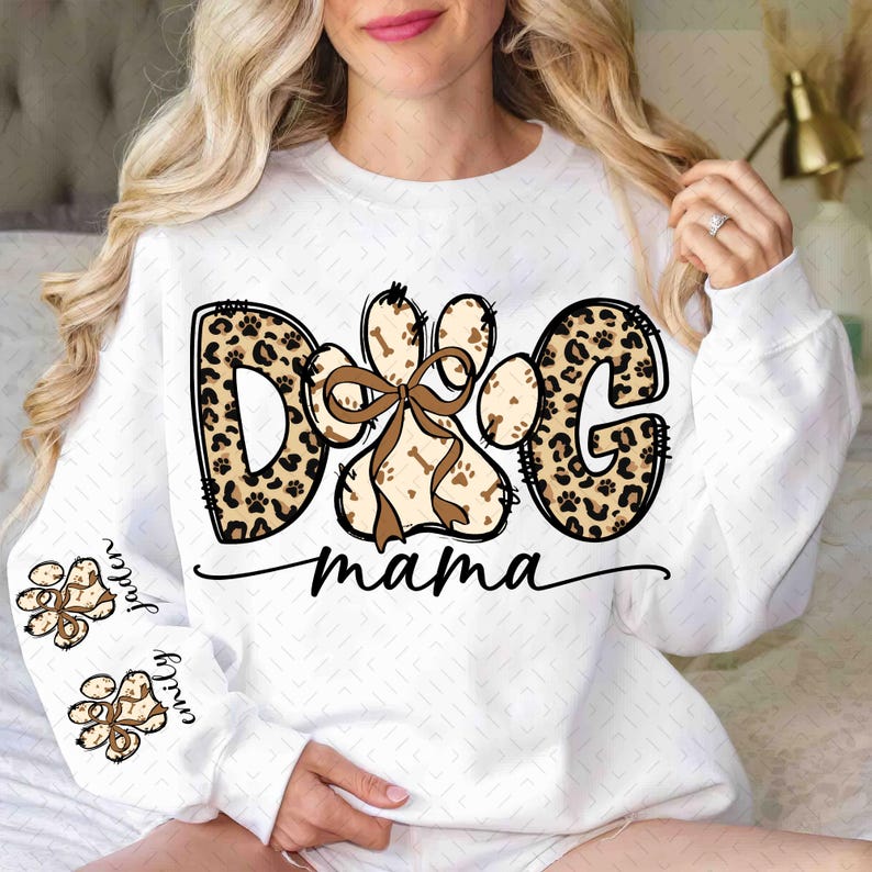 Puede incluir: Sudadera blanca con un dise&ntilde;o de "Dog Mama". El dise&ntilde;o presenta la palabra "DOG" en estampado de leopardo, una huella de pata con un lazo marr&oacute;n y la palabra "mama" en una fuente cursiva. Las mangas tienen huellas de patas con nombres.