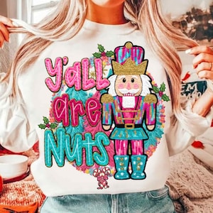 May include: White sweatshirt with a colorful graphic featuring a nutcracker soldier and the text "Y'all are Nuts" in pink and teal. The design includes holly and candy cane accents, perfect for the holiday season.