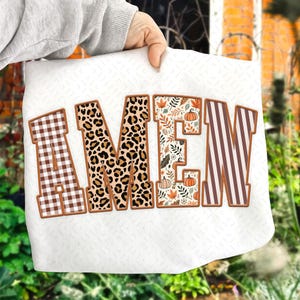 Puede incluir: Cojín blanco con la palabra "AMEN" bordada en marrón. Las letras tienen diferentes patrones: cuadros, estampado de leopardo, hojas de otoño y rayas. El cojín está sostenido por una persona.