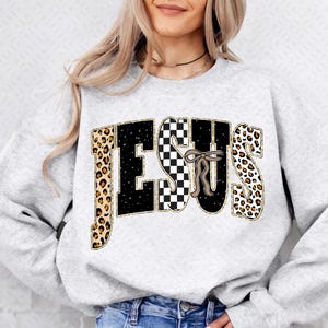 Könnte beinhalten: Ein hellgraues Sweatshirt mit dem Wort "JESUS" in einem auffälligen Design. Die Buchstaben haben einen Mix aus Leopardenmuster, schwarz-weißem Schachbrettmuster und einer Schleife. Das Sweatshirt ist ein lässiger, bequemer Stil.