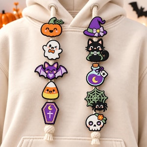 Puede incluir: Una sudadera con capucha de color beige con dos cordones blancos gruesos decorados con diez dijes de goma coloridos. Los dijes incluyen una calabaza, un fantasma, un murciélago, un dulce, un ataúd, un sombrero, un gato, una poción, una telaraña y una calavera.
