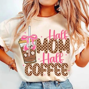 K&ouml;nnte beinhalten: Wei&szlig;es T-Shirt mit einem braunen und wei&szlig;en Karomuster und rosa Schriftzug "Half Mom Half Coffee". Auf der linken Seite des Shirts befinden sich ein rosa Band und ein braunes, eisgek&uuml;hltes Getr&auml;nk mit Strohhalm.