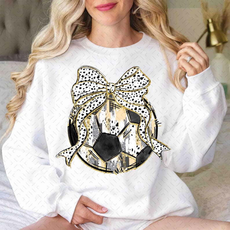 Op de afbeelding: Wit sweatshirt met een zwart-witte voetbal met een gouden en zwarte strik erop. De voetbal heeft een verweerde look met gouden accenten.